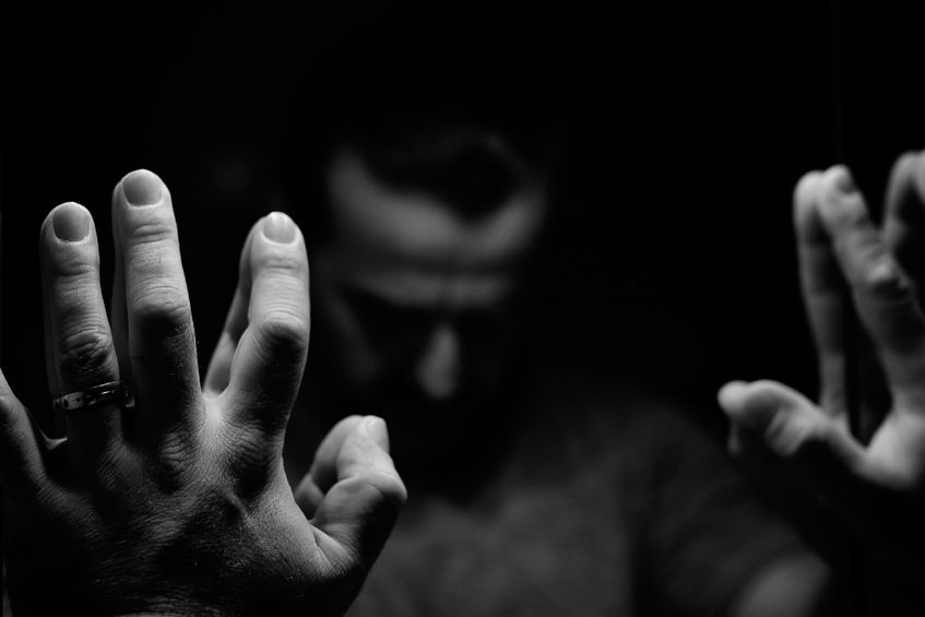 man in despair with raised hands and bowed hand, monochromatic image in a low light room looking in front of mirror
