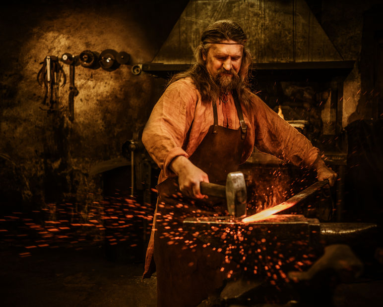 Senior blacksmith forging the molten metal on the anvil in smithy.